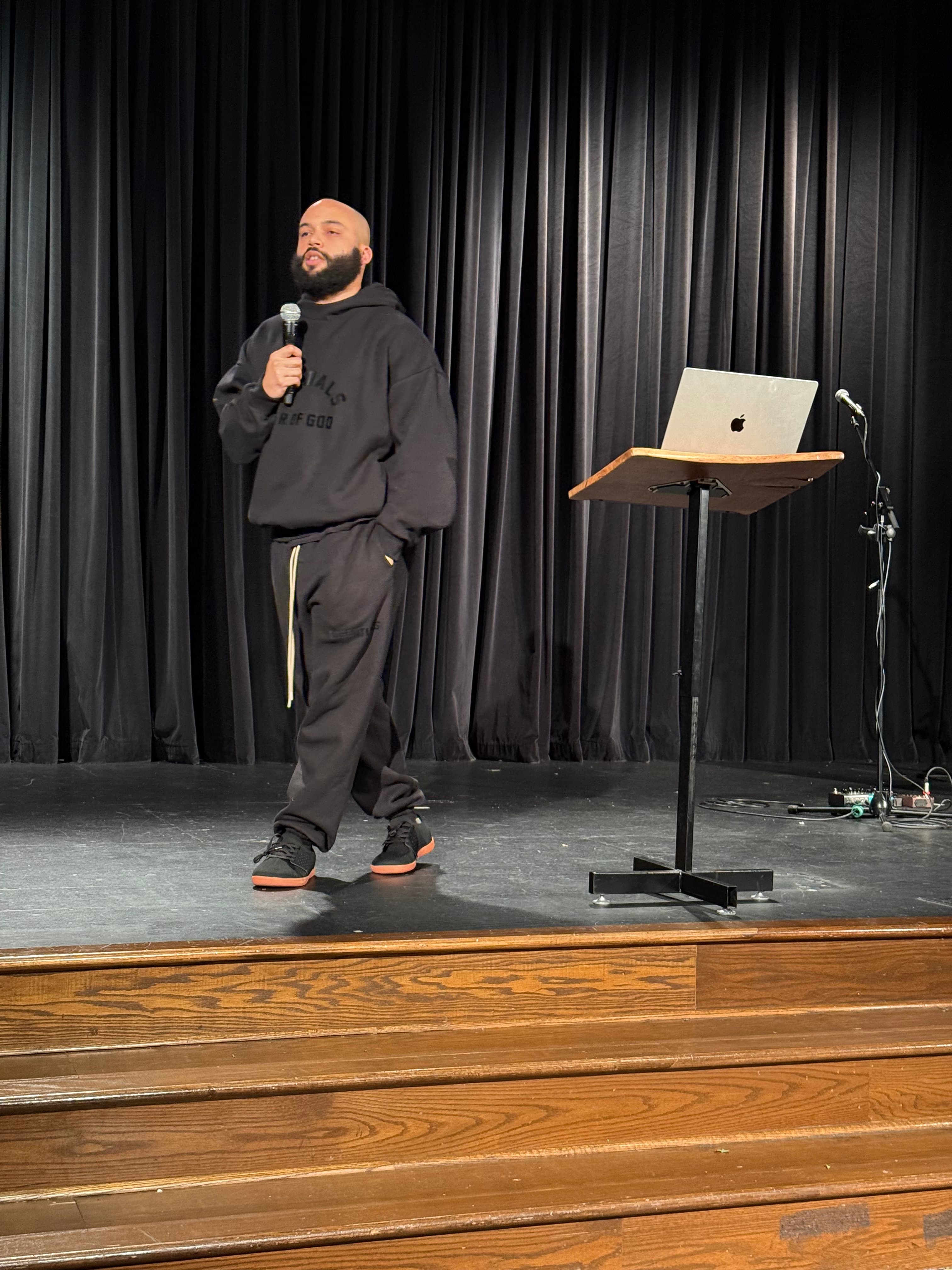 Blake M. Jones in a hoodie speaking on a dim stage, wireless microphone in hand