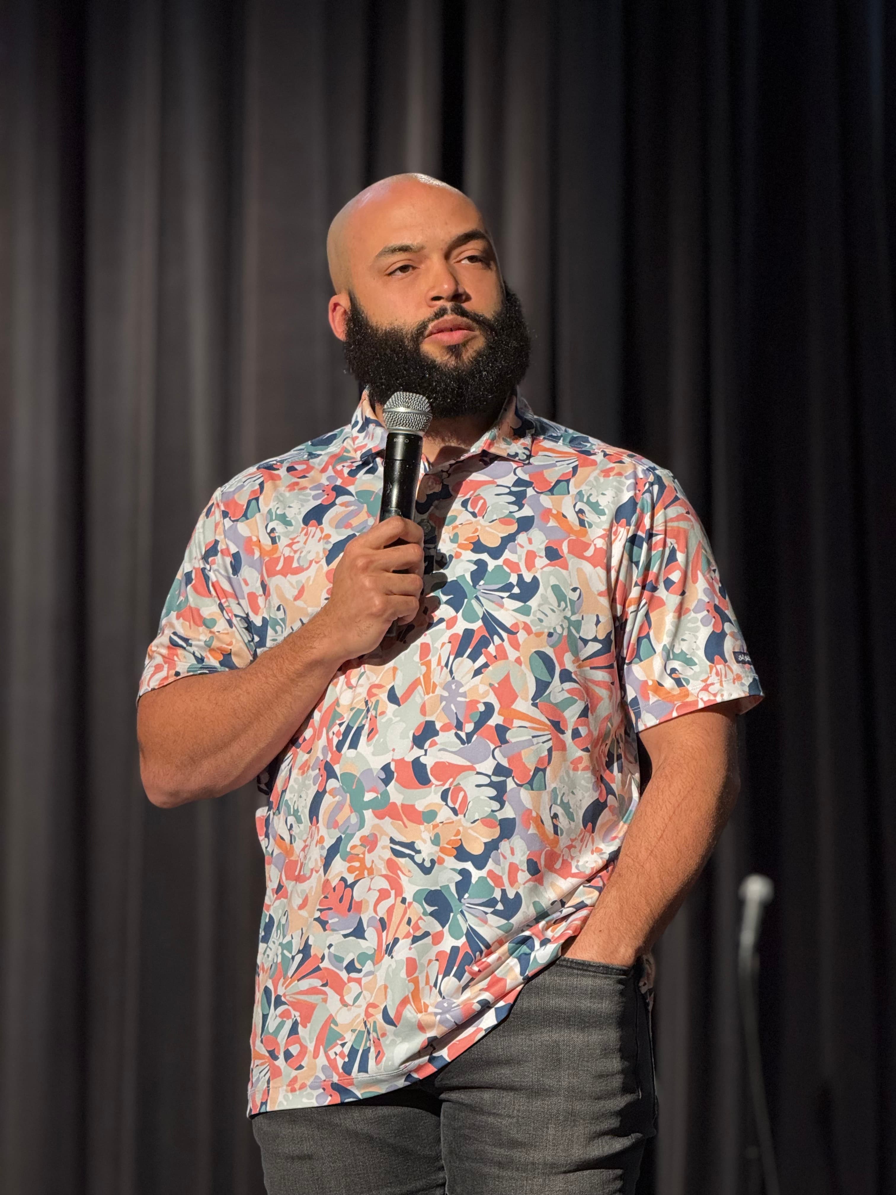 Blake M. Jones preaching with a handheld microphone, floral shirt against a dark stage curtain